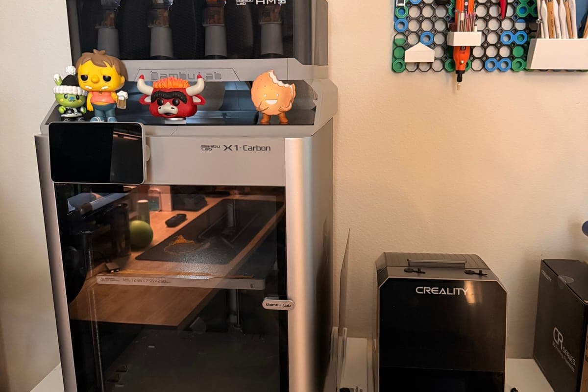 Bambu Lab X1-Carbon with an AMS on top, vinyl figures lined up on the deck, filament dryer alongside