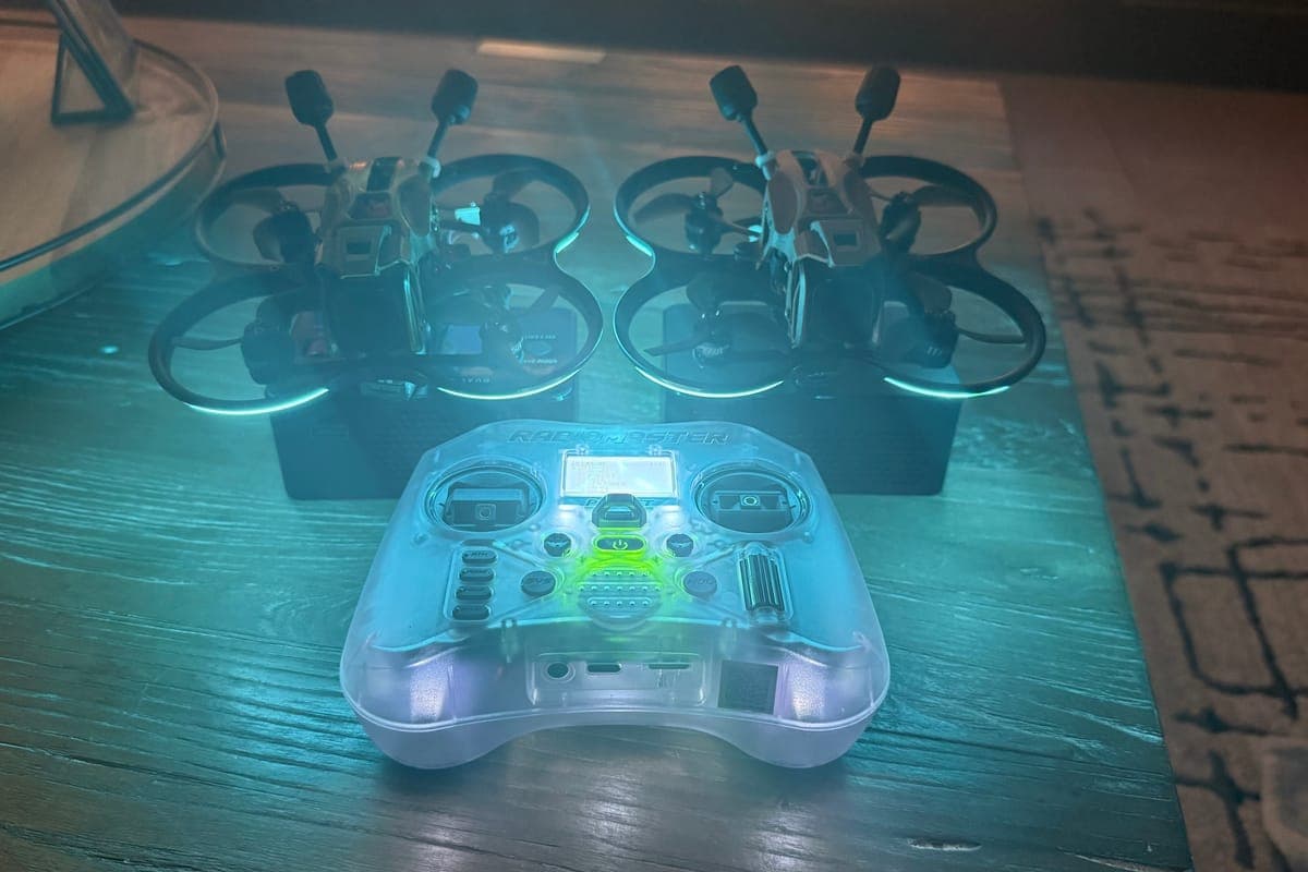 Two FPV drones on charging cases behind a translucent RadioMaster controller on a coffee table