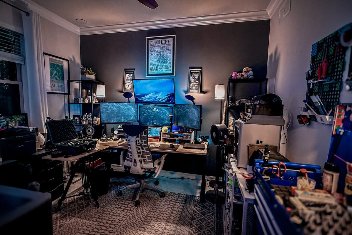 Home studio at dusk: triple monitors, a camera case on a side table, shelves and tool storage on both walls