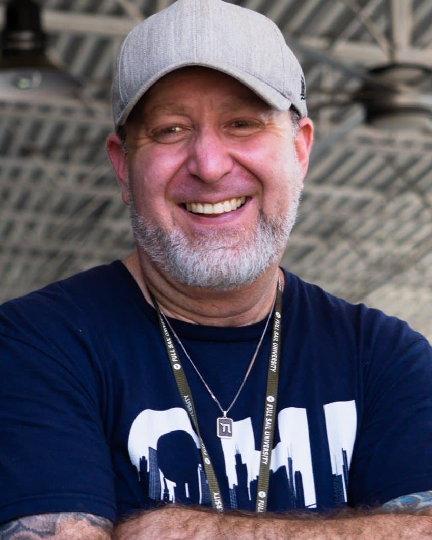 Michael smiling with arms crossed, in a gray cap and a Chicago skyline T-shirt, under an open-air pavilion.